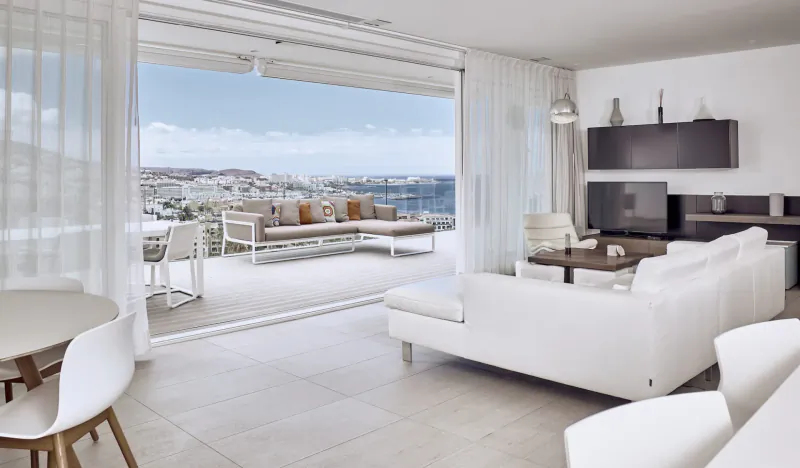 Spacious white modern living room in Baobab Suites, Tenerife, with open sliding doors to terrace overlooking sea and town.