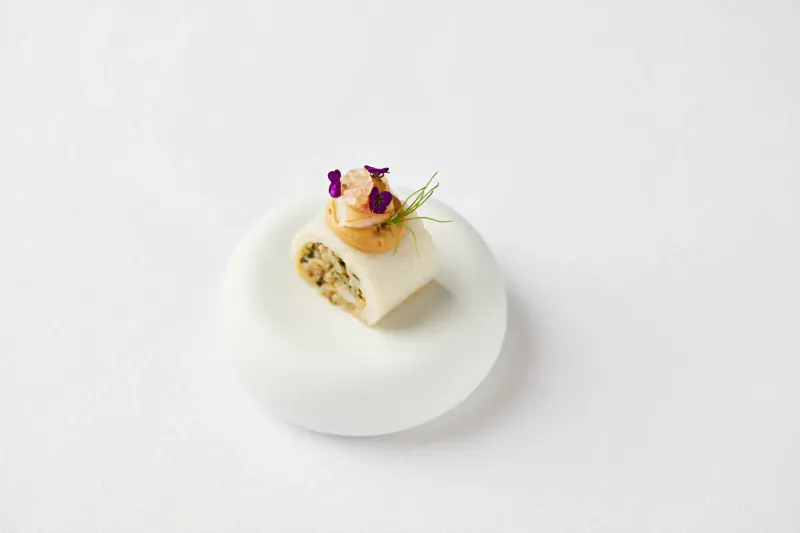 Close-up of gourmet white roll with green filling, pink prawn, purple flowers, and microgreens on white plate