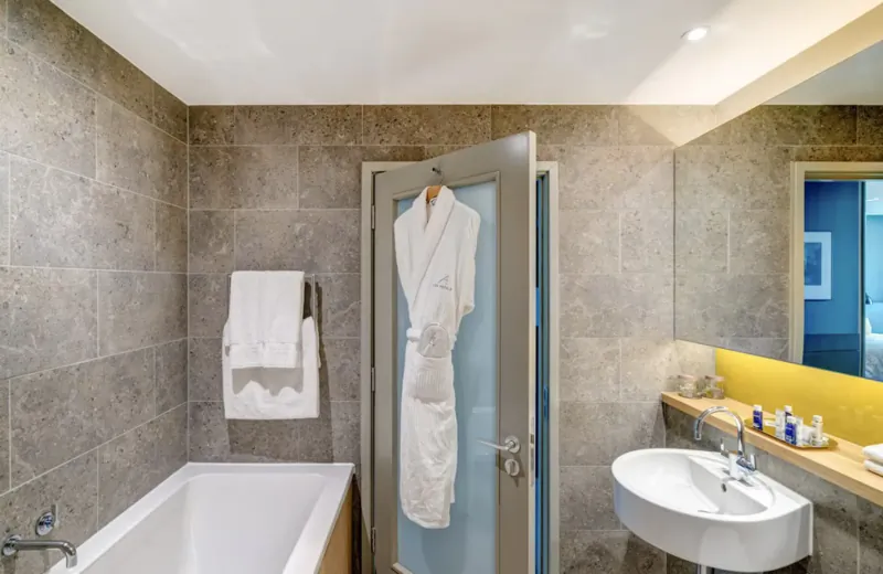 Modern hotel bathroom with white robe on open door, bathtub, white sink, towels, and toiletries.