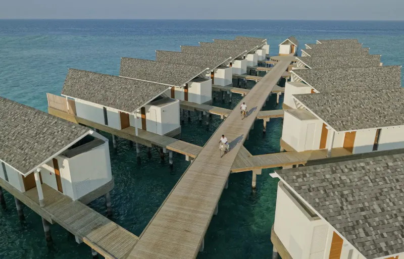 Aerial view of white overwater villas connected by wooden walkways over turquoise Maldives lagoon, with people walking.
