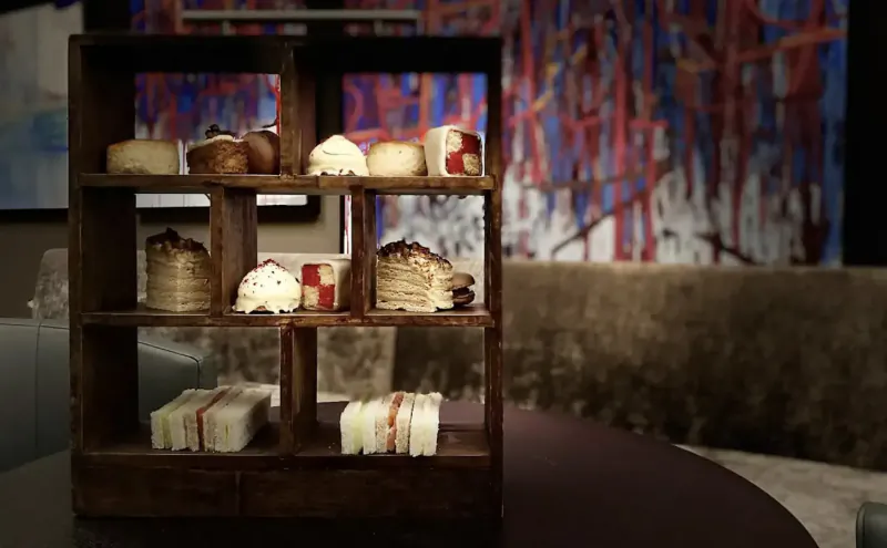 Wooden display shelf with assorted cakes and sandwiches in Twr y Felin hotel gallery, vibrant purple abstract background.