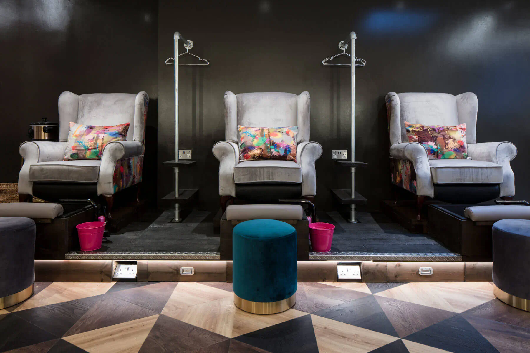 Three white pedicure chairs with colorful pillows in modern spa, foot baths, stools, and coat racks on dark wall