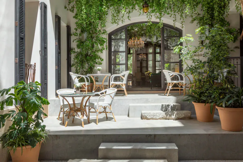 Elegant outdoor patio at Can Bordoy Grand House & Garden with white rattan chairs around glass table, lush hanging vines, potted plants, and arched doorway.