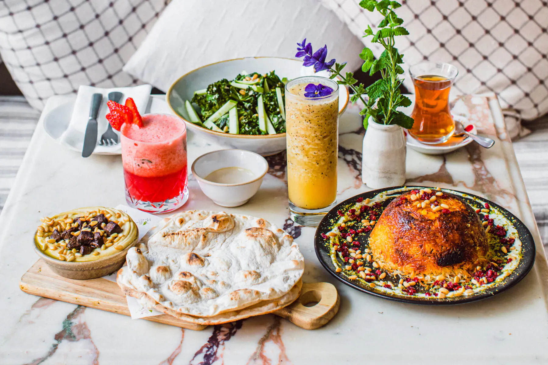 Assortment of Middle Eastern dishes on marble table: tahdig, flatbread, salads, dips, pink smoothie, orange juice, tea