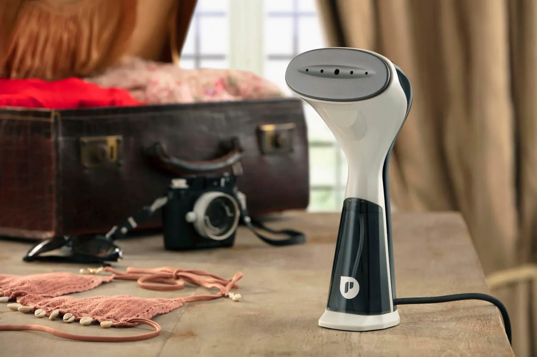 White and black Propress handheld steamer on wooden table with open vintage suitcase, red clothes, pink bikini, and retro camera.