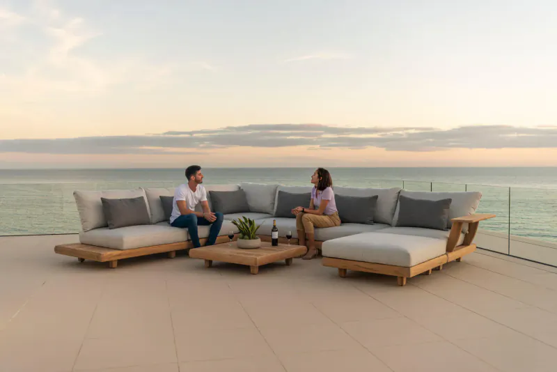 Man and woman chatting on Alexander Rose wooden outdoor L-shaped sofa on ocean-view terrace at sunset