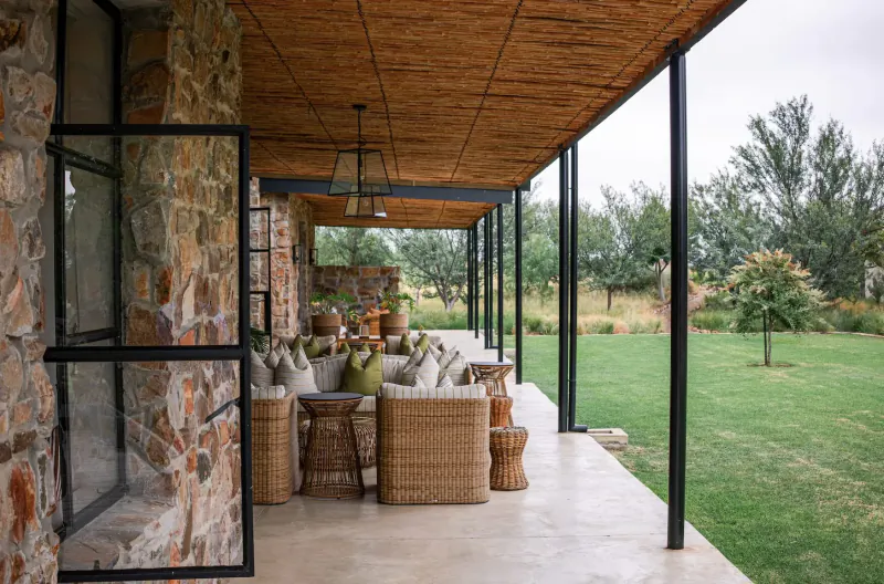 Stone and thatch veranda with rattan furniture, cushions, and lush green garden view at Simbavati Cederberg Ridge retreat.