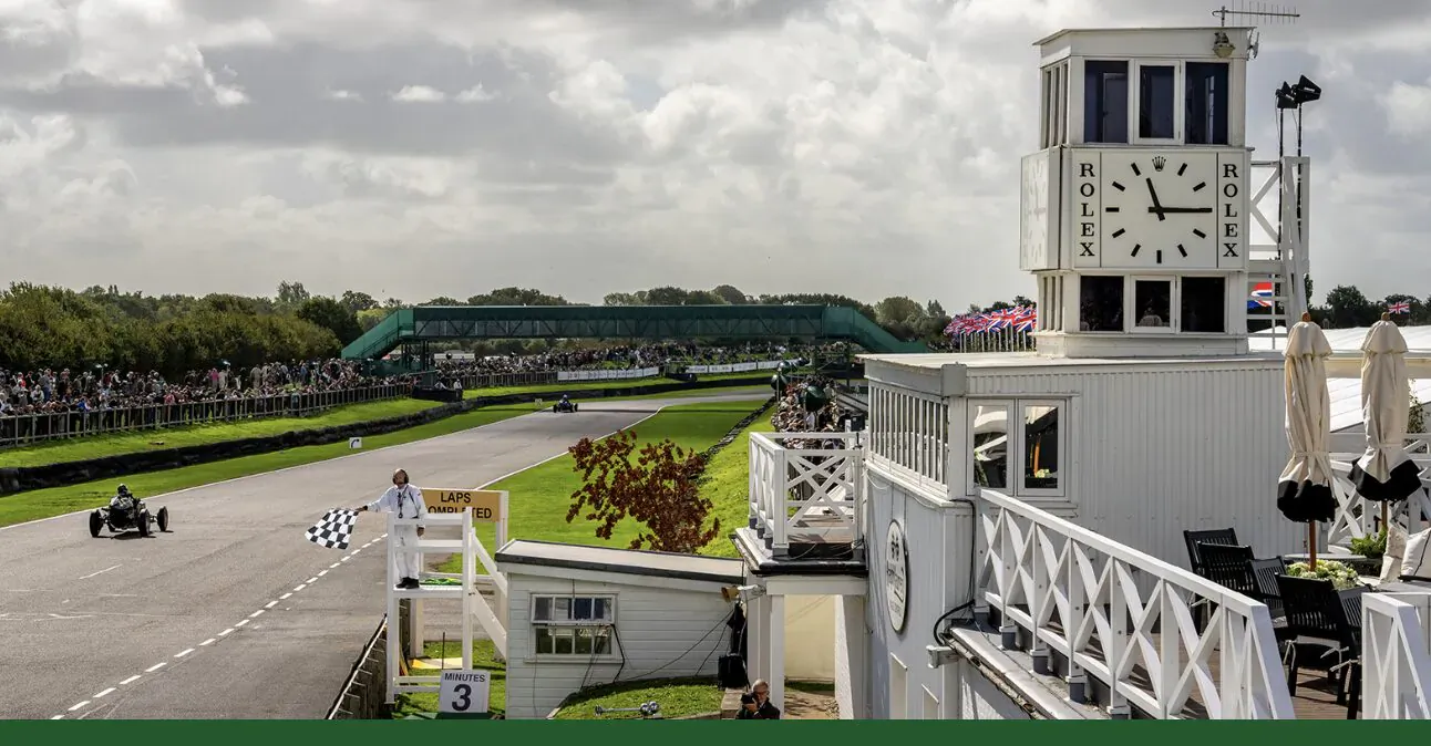 Single-seater race car speeds over checkered flag at Goodwood circuit towards white clock tower podium amid spectators.