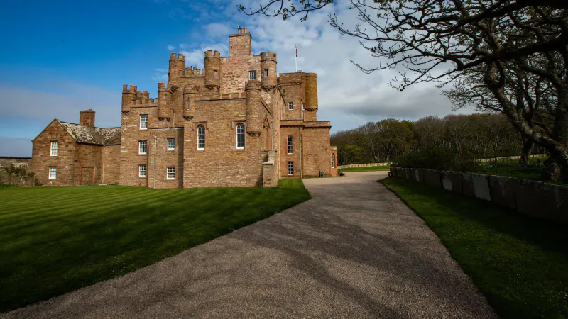 Red sandstone castle on North Coast 500, with driveway, green lawns, trees, and blue sky.
