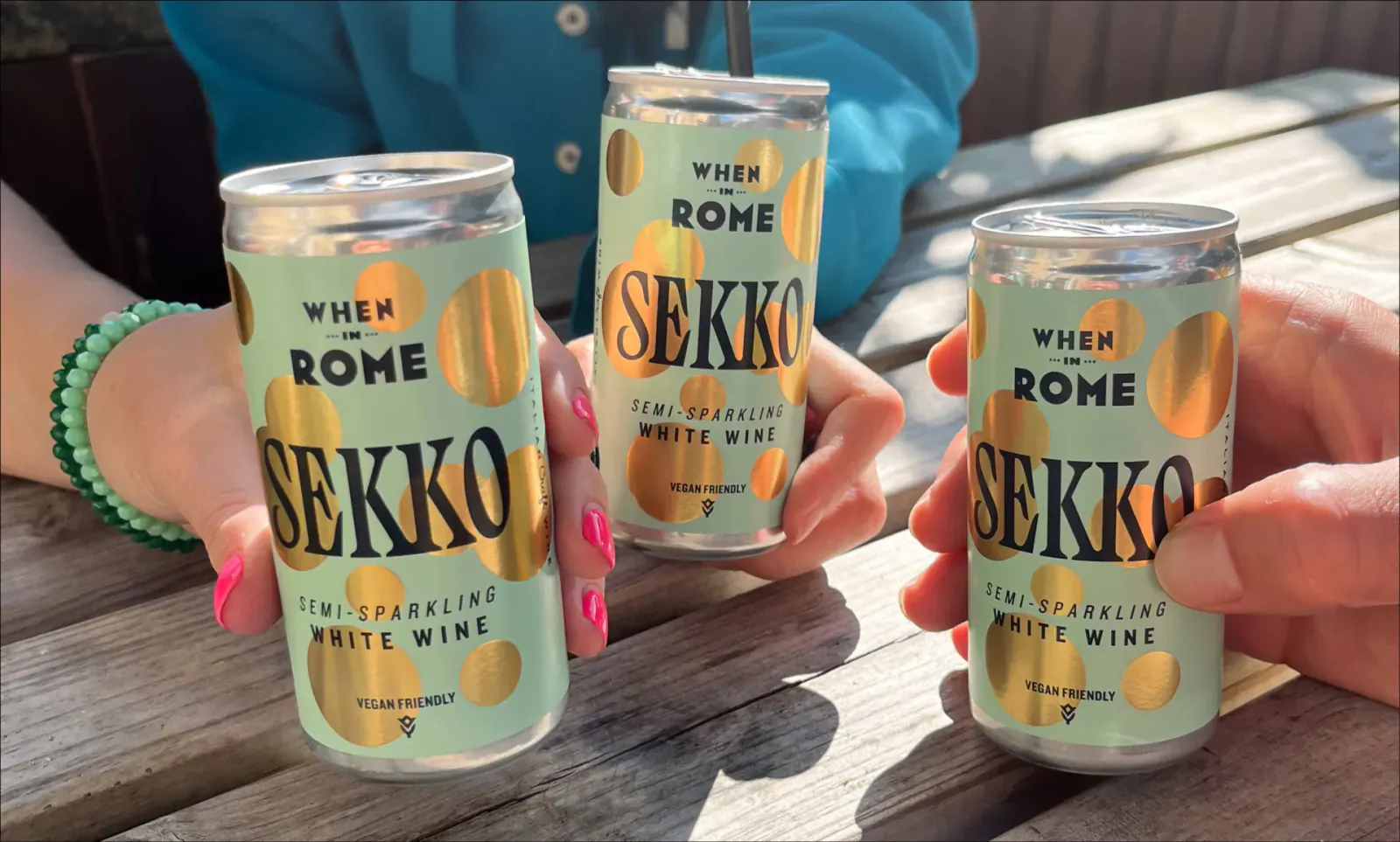 Hands holding three green Sekko white sparkling wine cans on a table, labeled 'White Wine'.