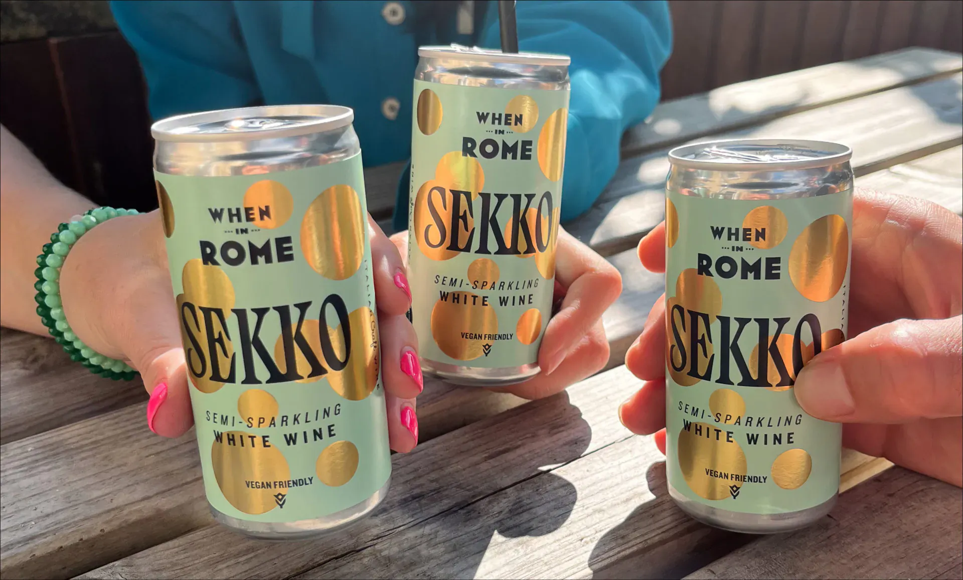 Hands holding three green Sekko white sparkling wine cans on a table, labeled 'White Wine'.