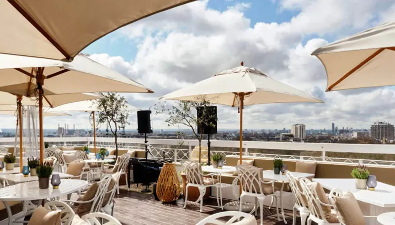 Rooftop terrace at The Dorchester Hotel with white umbrellas, wicker chairs, tables, speakers, and London skyline view.