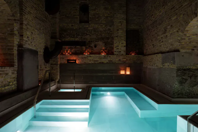 Candle-lit indoor pool in ancient brick-walled bathhouse at Aire Ancient Baths, glowing turquoise water and steps.