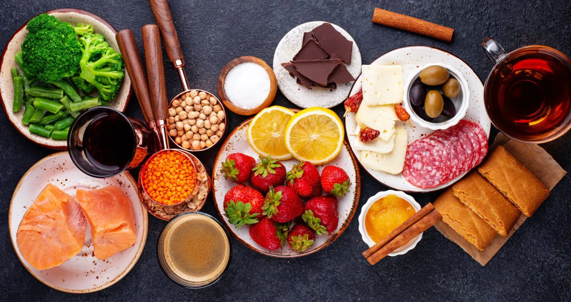 Assortment of healthy foods on dark surface: broccoli, salmon, lentils, strawberries, cheese, olives, chocolate, tea, wine.