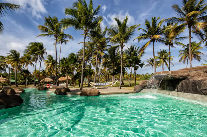Tropical resort pool with turquoise water, hammock between palms, waterfall, and thatched huts at Palm Island.