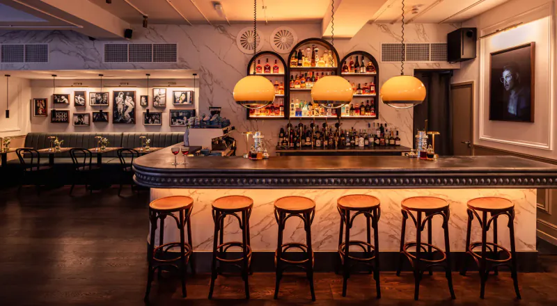 Elegant Ziggy Green bar in London with curved counter, glowing pendant lamps, backlit bottles, stools, and art on walls.