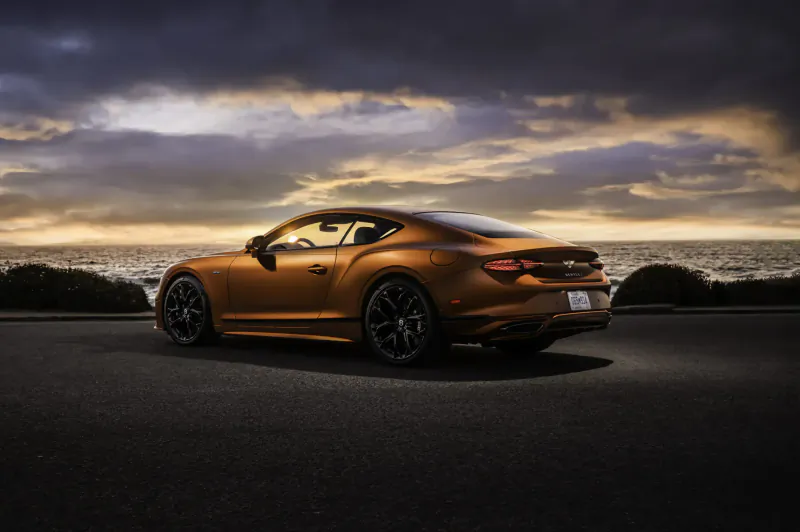 Rear view of orange Bentley Continental GT Speed coupe parked on road by ocean at sunset with dramatic clouds.