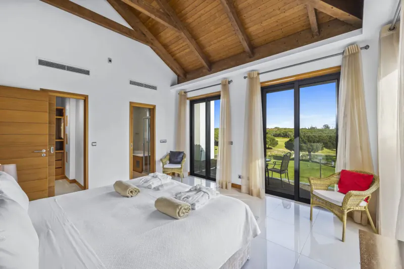 Spacious bedroom in Vale do Lobo resort with wooden beams, white bed with towels, chairs, and sliding doors to golf course view.
