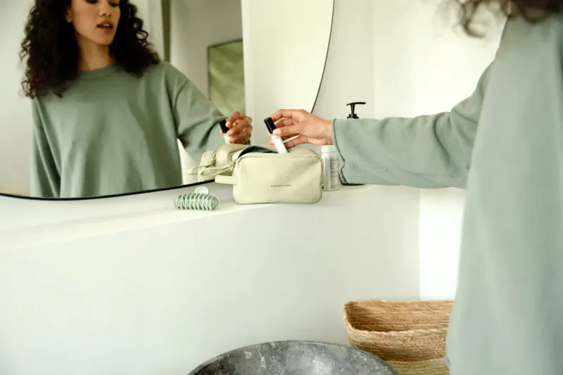 Woman in green sweater holding black item over sink in stylish bathroom mirror reflection, with vanity bag and basket.