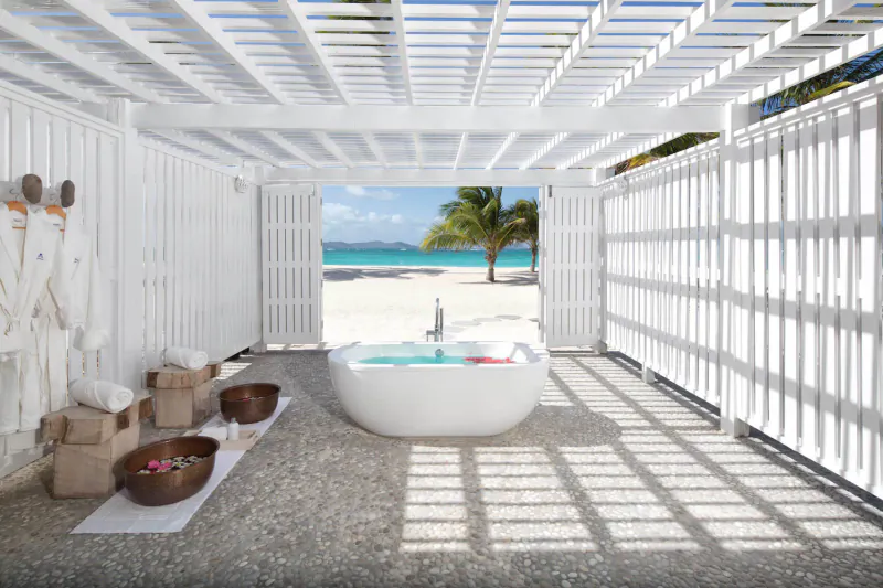 Open white lattice pergola at Palm Island Resort spa with freestanding tub, turquoise water, beach, and palm trees.