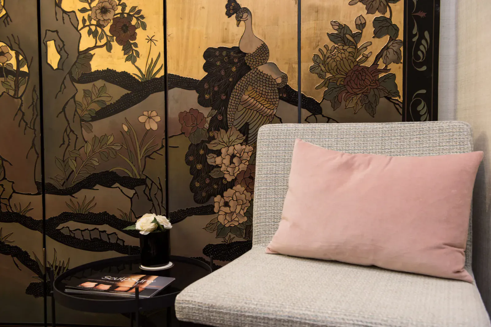 Cozy clinic corner with gray armchair, pink pillow, small table with white flowers, and ornate golden peacock screen.