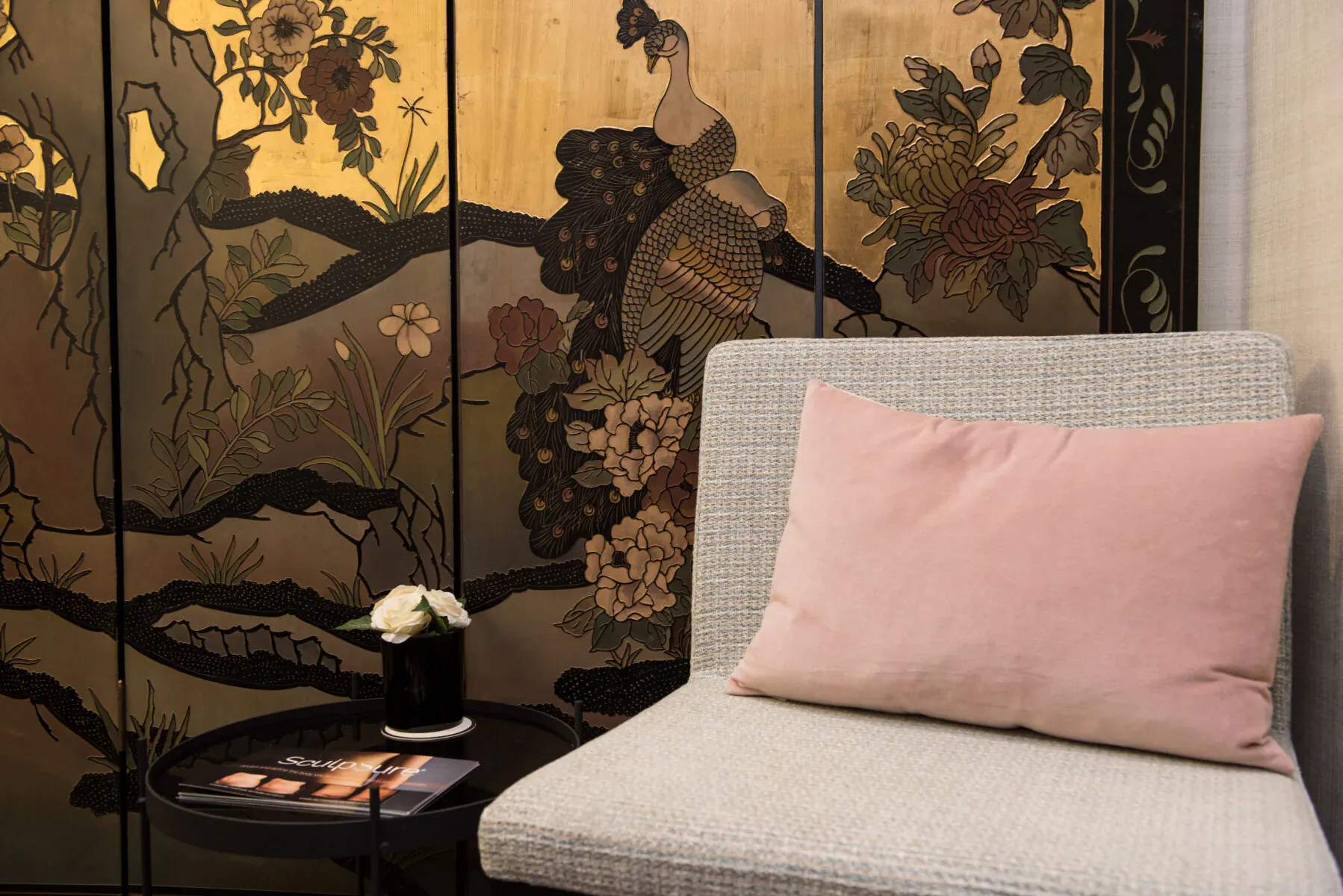 Cozy clinic corner with gray armchair, pink pillow, small table with white flowers, and ornate golden peacock screen.