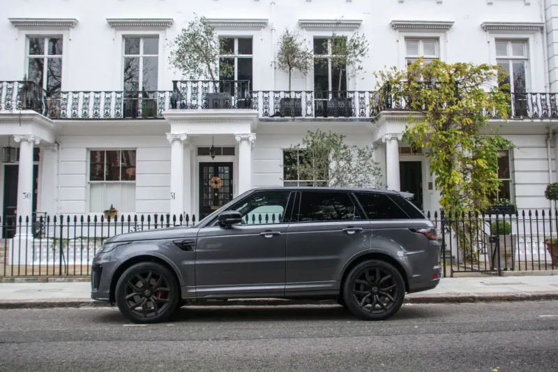 Gray Range Rover parked on street in front of elegant white terraced townhouses with plants and railings.
