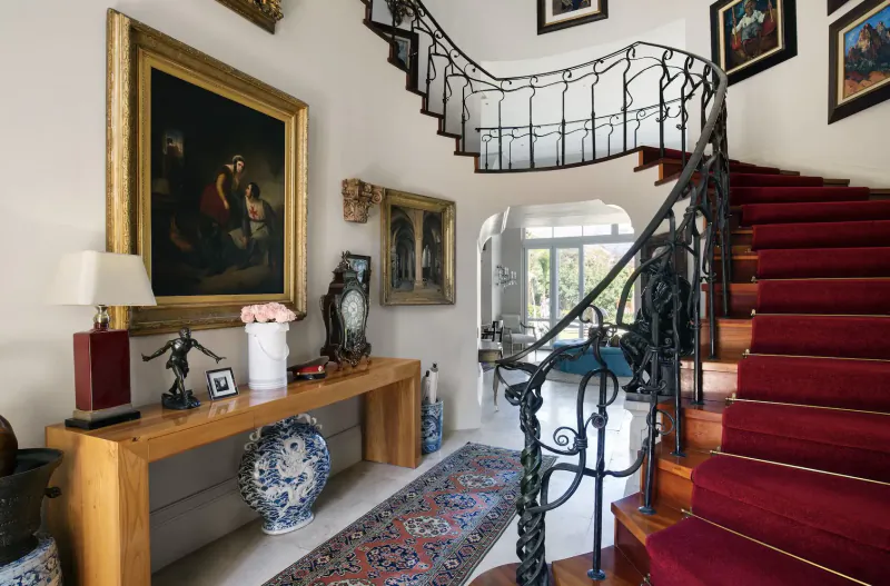 Elegant entryway of 21 Nettleton in Clifton, Cape Town, with ornate wrought-iron curved red-carpeted staircase, gold-framed paintings, console table with vase and lamp.