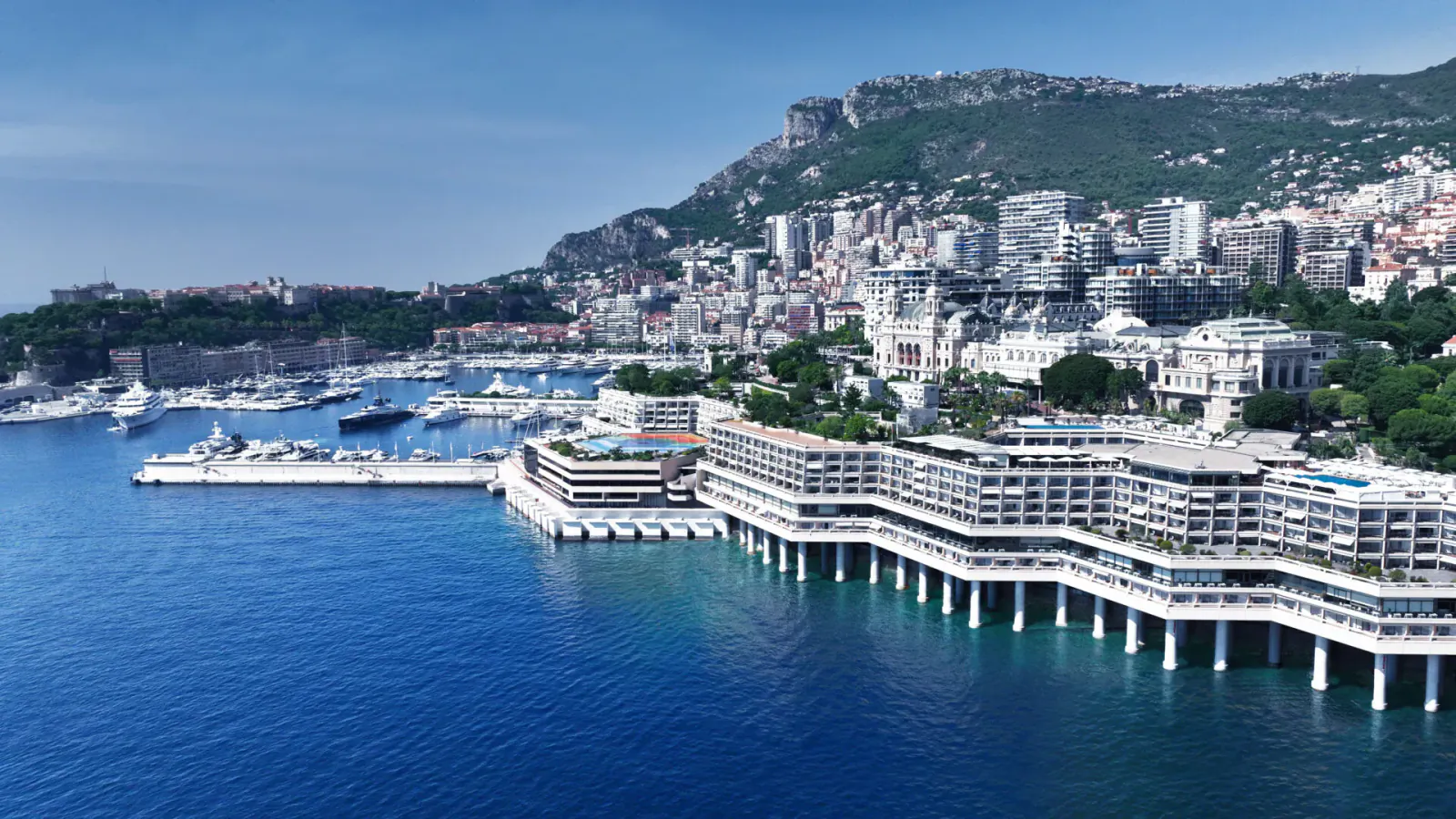 Aerial view of Fairmont Monte Carlo hotel on cliffs above marina with yachts, celebrating 50th anniversary renovation.