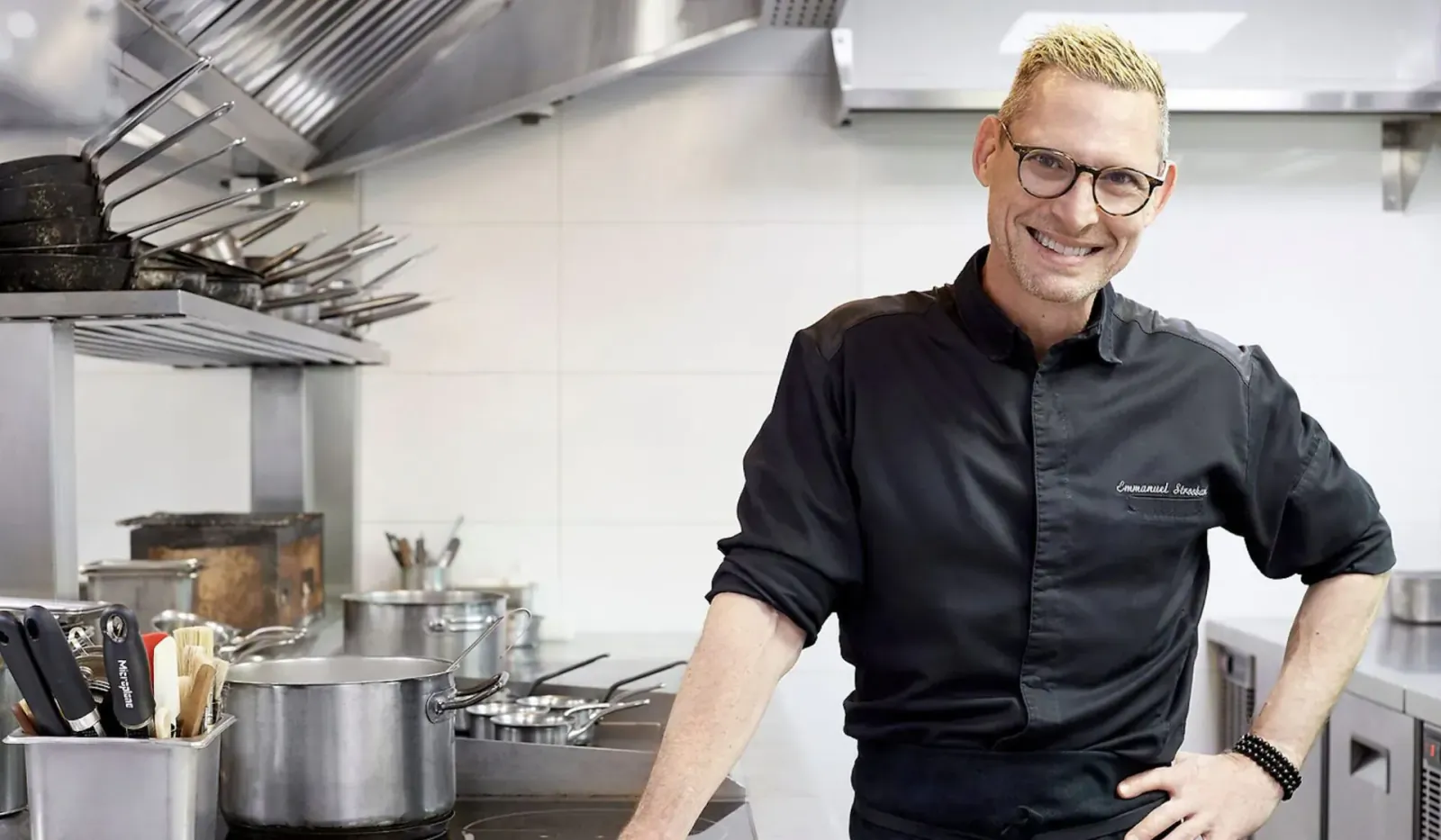 Smiling chef with blond hair and glasses in black uniform, standing confidently in professional kitchen at Saint Pierre.