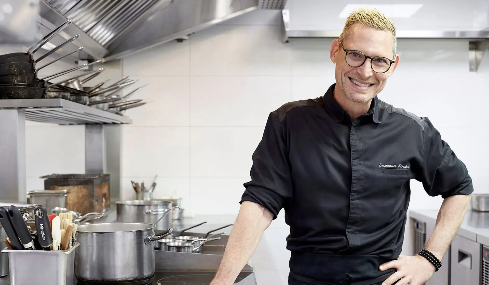 Smiling chef with blond hair and glasses in black uniform, standing confidently in professional kitchen at Saint Pierre.