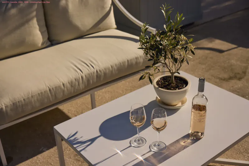 Outdoor white table with two glasses of white wine, rosé bottle, and potted olive tree on beige sofa, Léoube Estate.