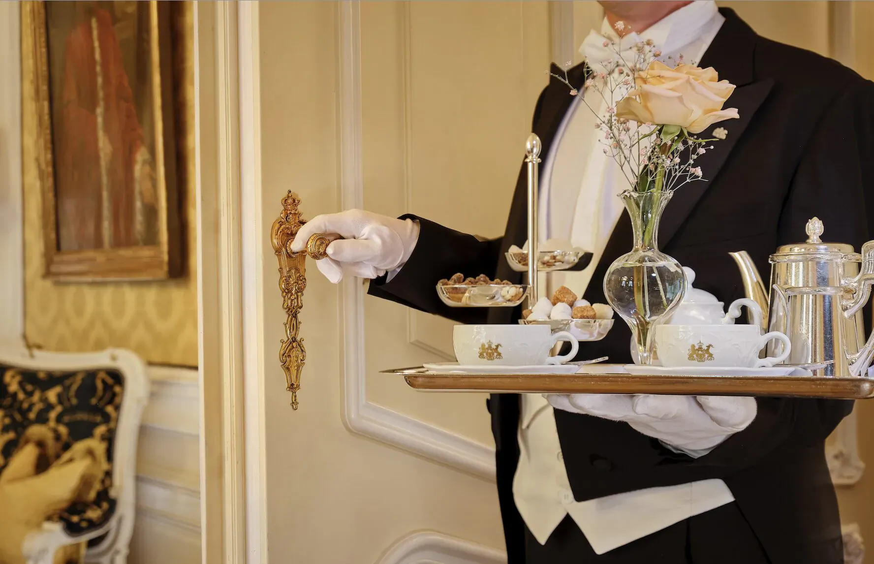 Tuxedoed waiter in white gloves holding silver tray with teapot, cups, and pastries in luxurious hotel doorway