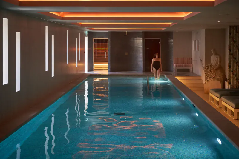 Woman in black swimsuit walking down steps into a stylish indoor pool at The NICI hotel, warm ambient lighting.