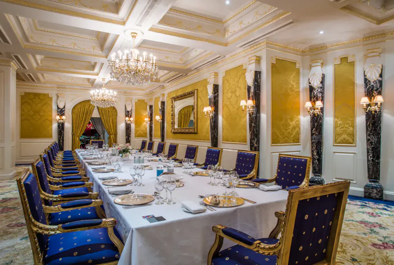 Elegant dining room at Bentley Hotel with long table set for meal, blue velvet chairs, chandeliers, gold walls.