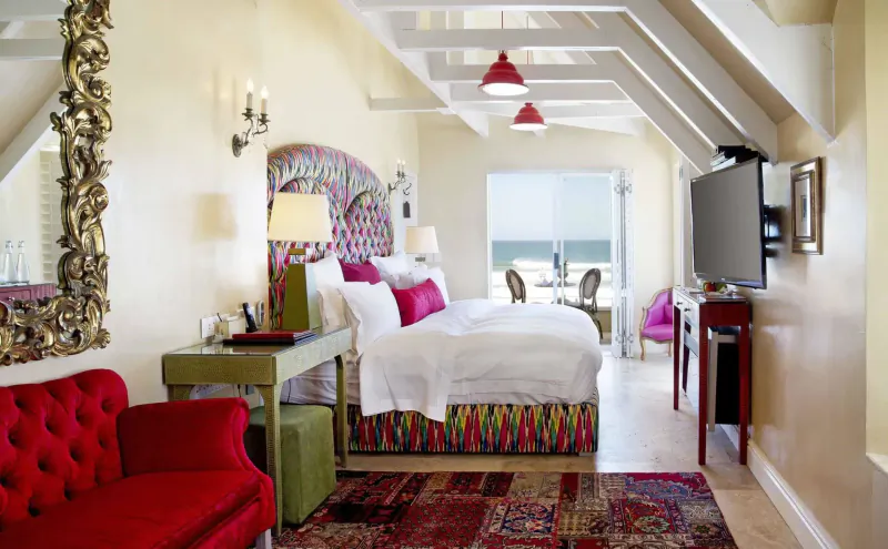 Luxurious attic hotel room with colorful patterned bed, red armchair, ocean view window, and ornate mirrors.