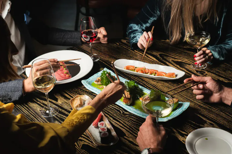 Group at wooden table in dim light, enjoying sushi, sashimi, greens, and wine glasses with chopsticks.