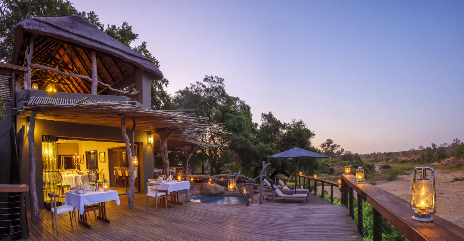 Twilight view of rustic savanna lodge deck with lantern-lit dining table, pool, loungers, and bushveld backdrop.