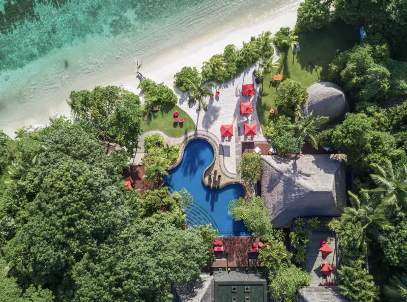 Aerial view of Anantara Maia Seychelles Villas' blue lagoon pool, red umbrellas, white beach, and turquoise ocean amid lush greenery.