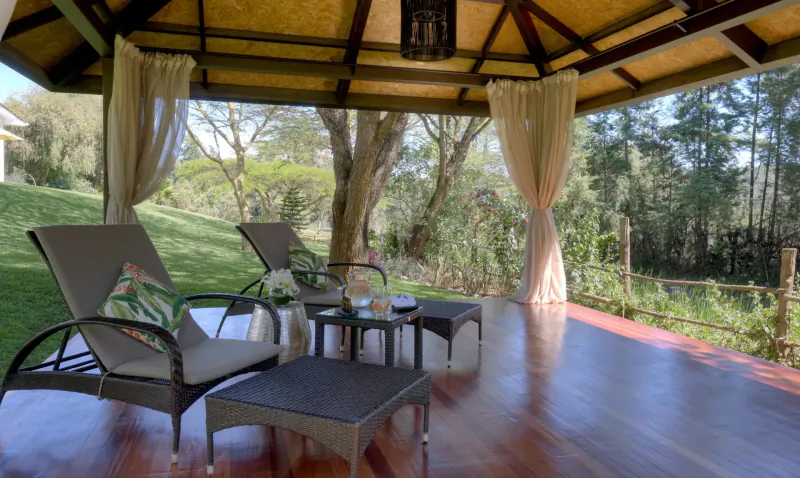 Luxurious outdoor deck with cushioned lounge chairs, low table, and sheer curtains under thatched pavilion overlooking lush green forest.