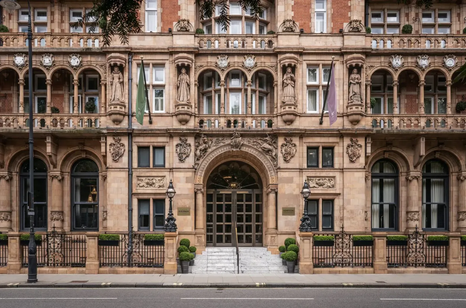 Grand entrance of Kimpton Fitzroy London hotel, ornate Victorian facade with green and purple flags, steps, and planters.