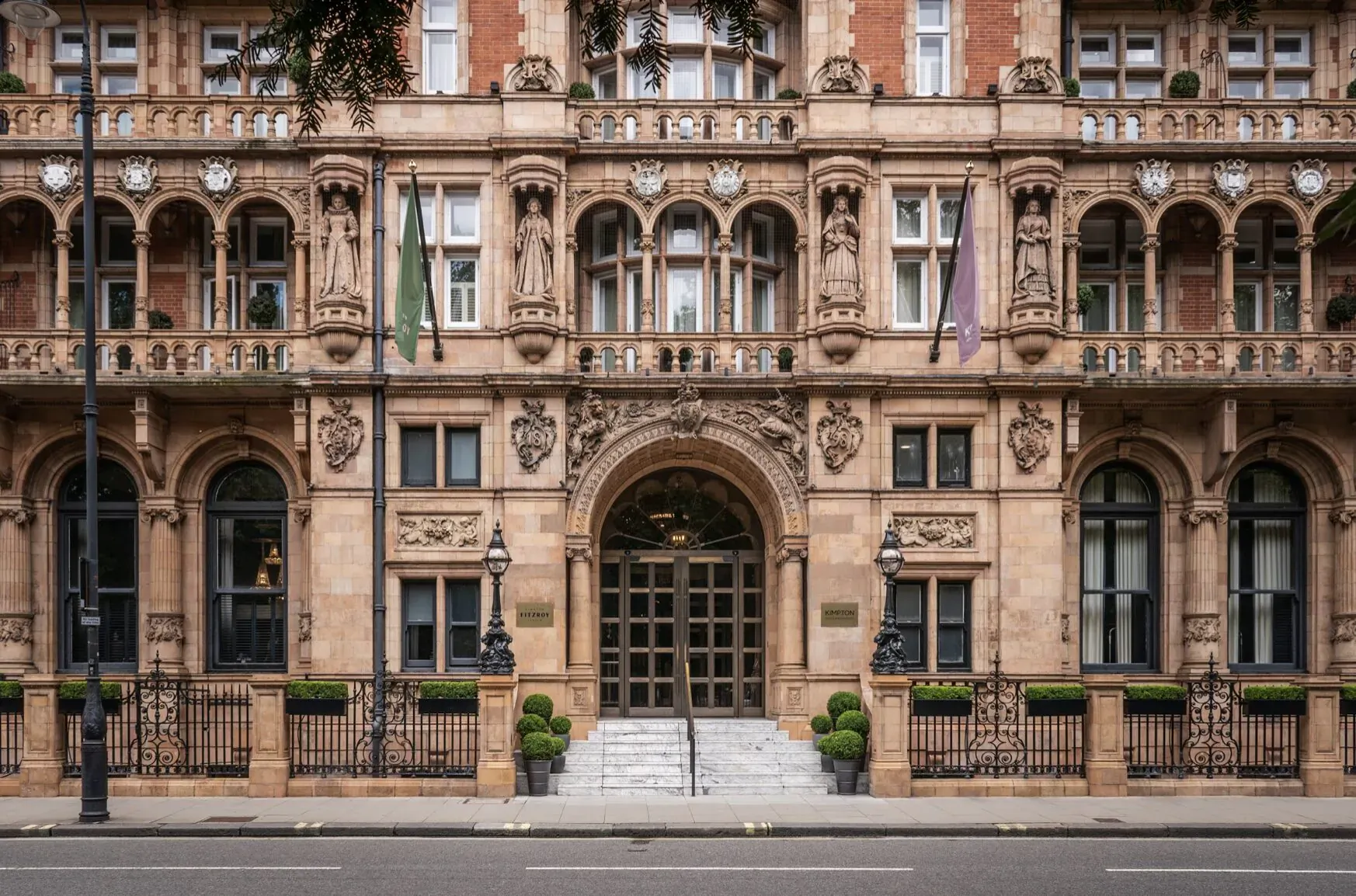Grand entrance of Kimpton Fitzroy London hotel, ornate Victorian facade with green and purple flags, steps, and planters.