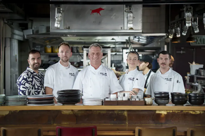 Group of chefs in white 'TPL' uniforms standing behind counter with bowls and plates in modern kitchen, red bull logo above.