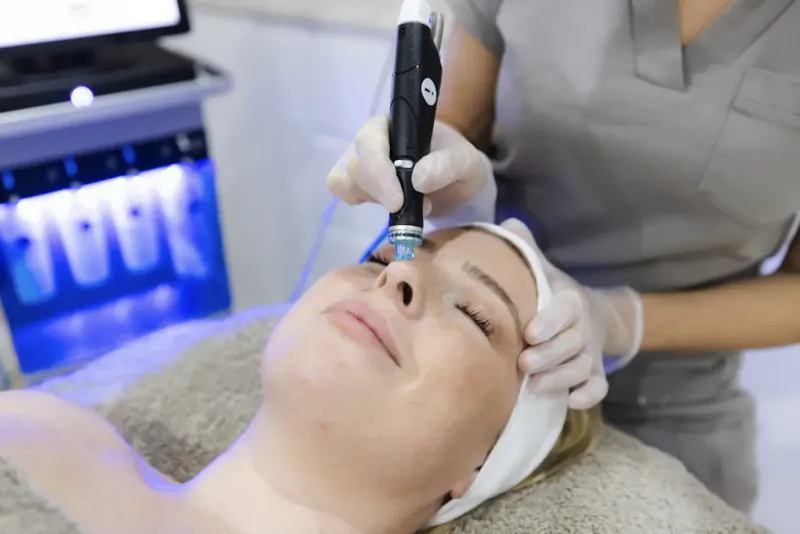 Nurse in scrubs performs microneedling facial treatment on relaxed female client lying on spa bed with blue LED lights.
