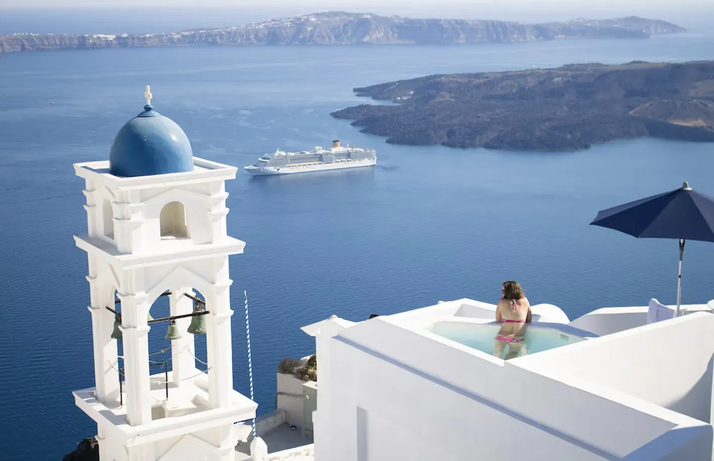 Woman in bikini in infinity pool by white Santorini bell tower, overlooking blue Aegean Sea, cruise ship, and islands