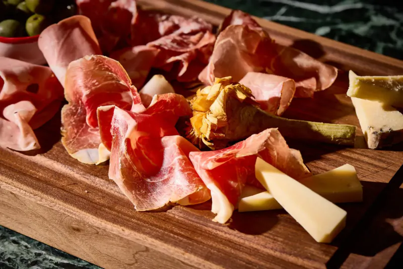 Charcuterie board with prosciutto, salami, cheese wedges, mustard, and olives on wooden board