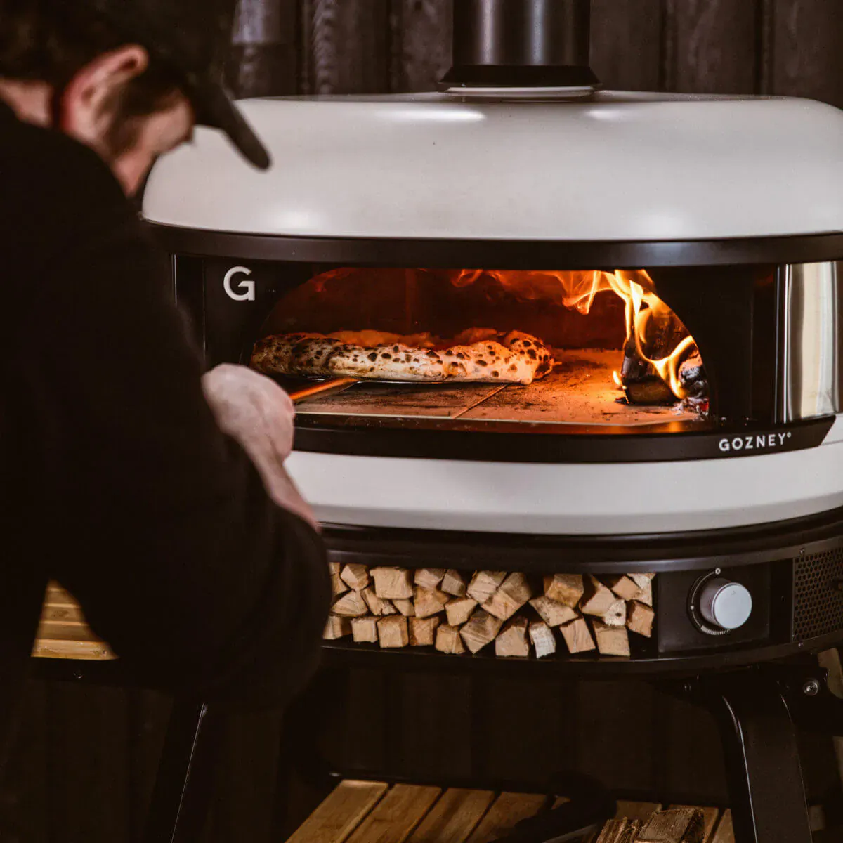 Man wearing cap checks pizzas baking in glowing Gozney wood-fired oven with firewood stack below.