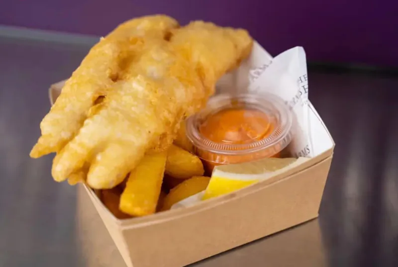 Two golden battered fish fillets, chips, orange sauce, and lemon wedge in a white-lined brown paper box.