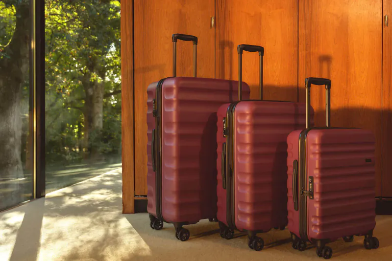 Three matching maroon hardshell suitcases from Antler Clifton Collection against wooden wall with garden view.
