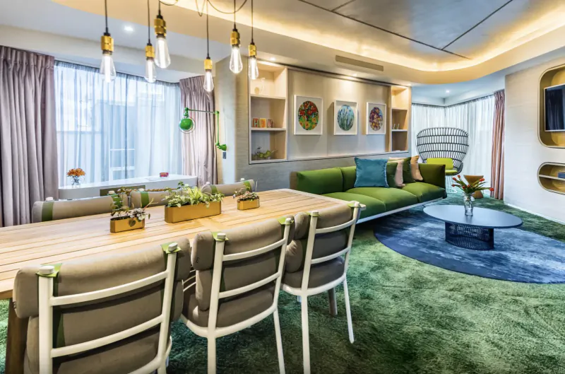 Modern hotel living room with green sofa, wooden dining table, pendant lights, plants, and city view windows.